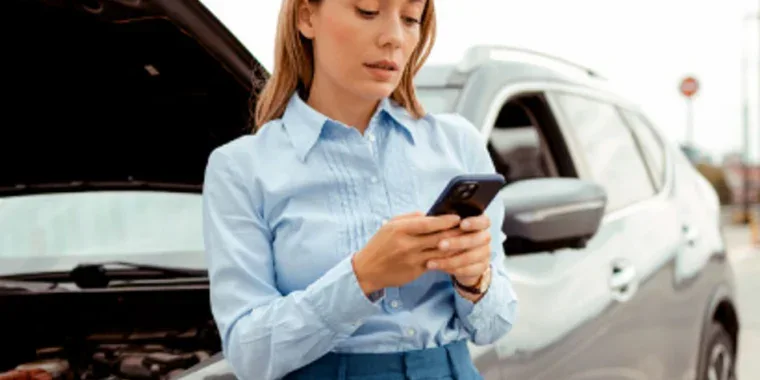 Woman standing by a car with the hood open, using her phone, possibly seeking assistance for a car breakdown.