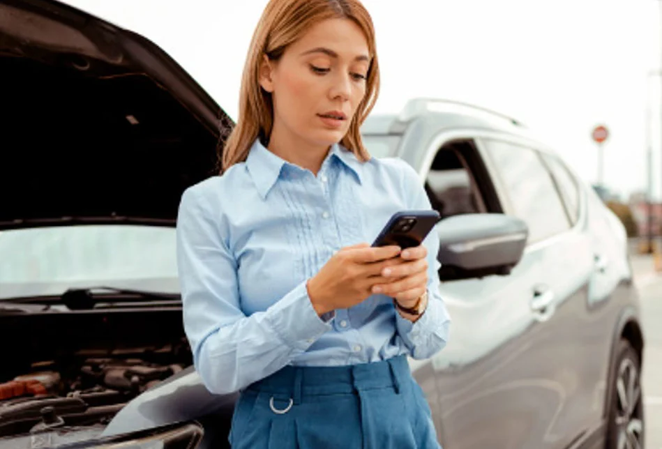 Woman standing by a car with the hood open, using her phone, possibly seeking assistance for a car breakdown.