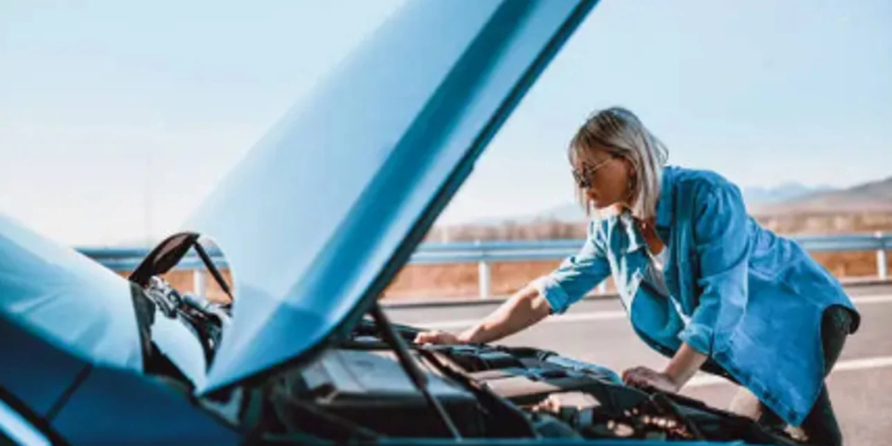 A person examines the engine under the raised hood of a car parked on the roadside during the day.