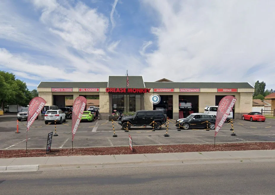 A Grease Monkey auto service center at 2100 Channing Way, Idaho Falls, the home of the 15 minute oil change with cars parked outside, red flags advertising services, and signage for oil change, alignment, maintenance, and repair—helping ensure engine longevity.
