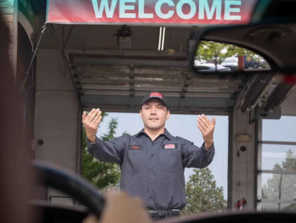 A car service worker in uniform stands under a "WELCOME" sign, directing a vehicle to move forward inside a service bay.