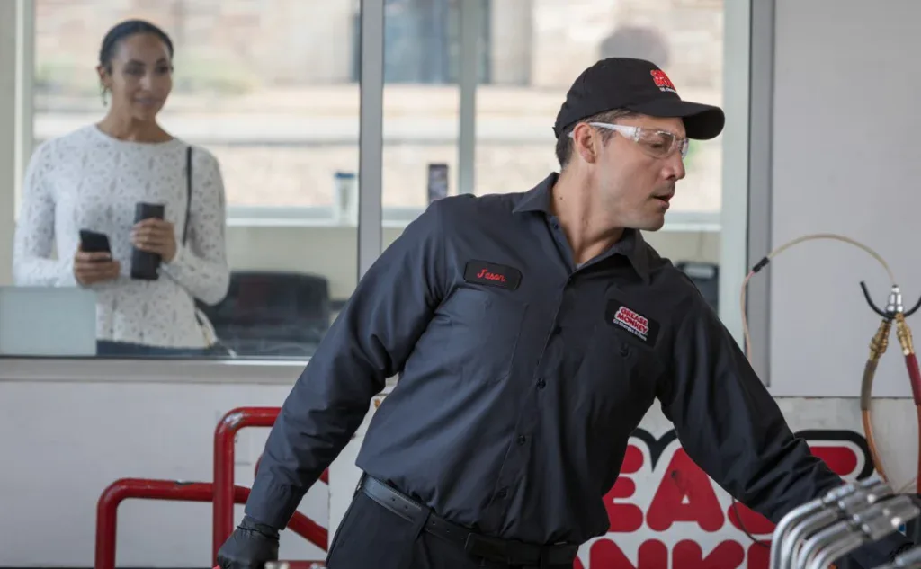 A mechanic in uniform works in a garage while a woman with a phone stands outside the window watching him.