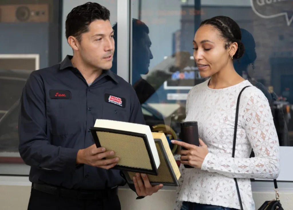 An auto mechanic shows two air filters to a customer standing outside a repair shop; the customer holds a coffee cup and looks at the filters.