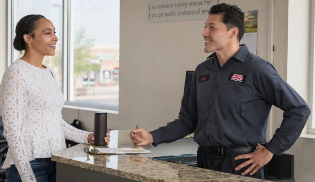 A woman stands at a service counter speaking with a male employee in a dark uniform who is holding a pen and clipboard.