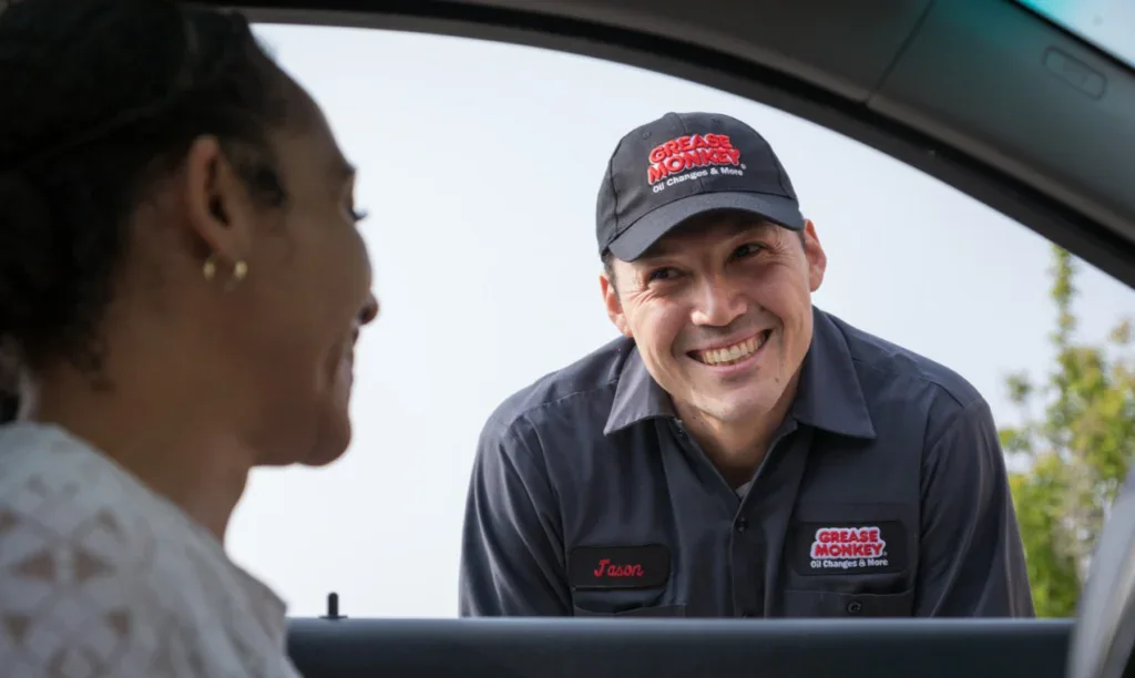 A Grease Monkey employee stands outside a car window, smiling and talking with a customer seated inside the vehicle.