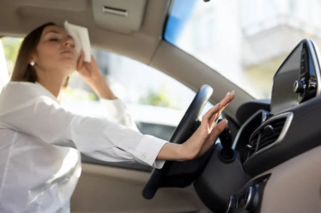 A person sits in a car in the 83440 zip code, wiping their forehead with a tissue and reaching to adjust the Rexburg vehicle AC controls on the dashboard.