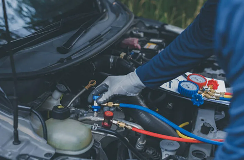 A mechanic wearing gloves is servicing a car engine with air conditioning gauges and hoses connected under the hood, ensuring expert AC care for optimal performance.