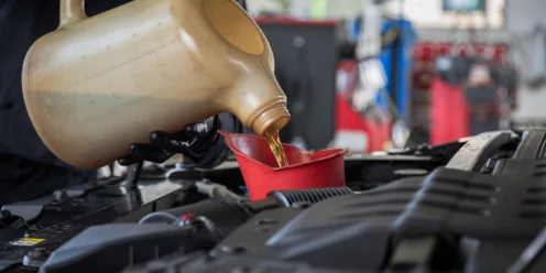 A person pours motor oil from a large container into a red funnel positioned in a car engine bay while performing engine services at an automotive service shop.