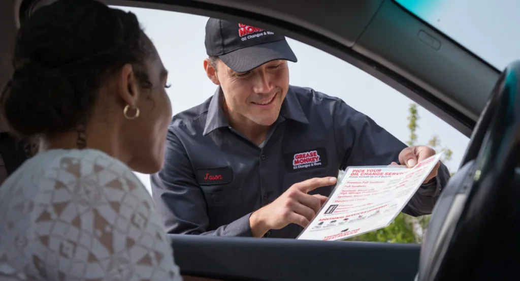 A technician in uniform shows a printed service menu to a woman sitting in her car, discussing options through the open car window.