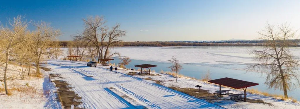 A snow-covered lakeside park with picnic shelters, bare trees, two residents, and a frozen lake under a clear blue sky.