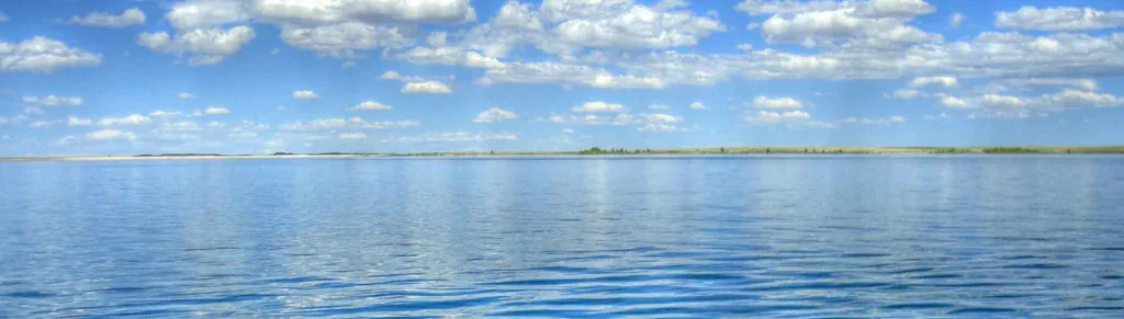A calm body of water under a blue sky with scattered white clouds, a distant, low shoreline on the horizon, and a faint hint of aurora shimmering above.