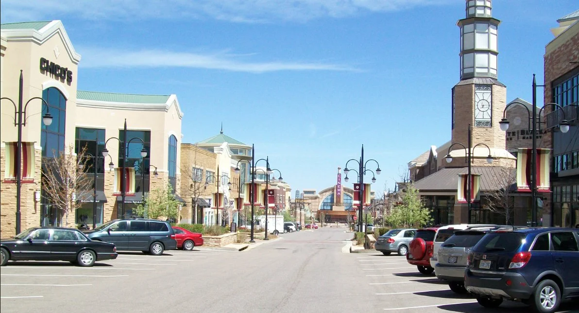 A wide street in a modern shopping center with parked cars on both sides, storefronts, street lamps, and a clock tower under a clear blue sky.