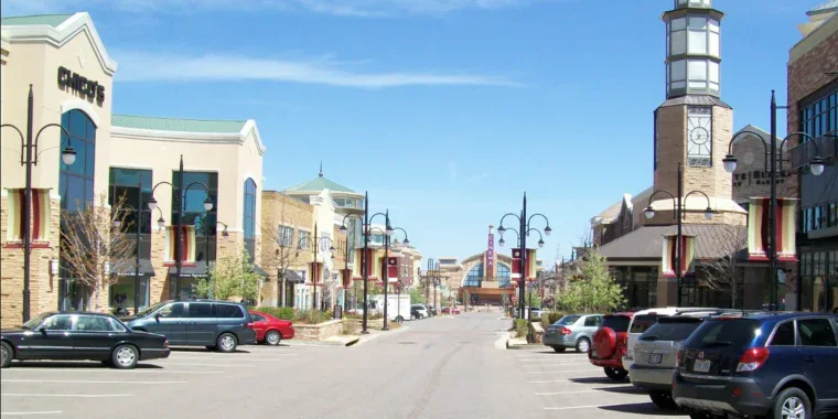 A wide street in a modern shopping center with parked cars on both sides, storefronts, street lamps, and a clock tower under a clear blue sky.