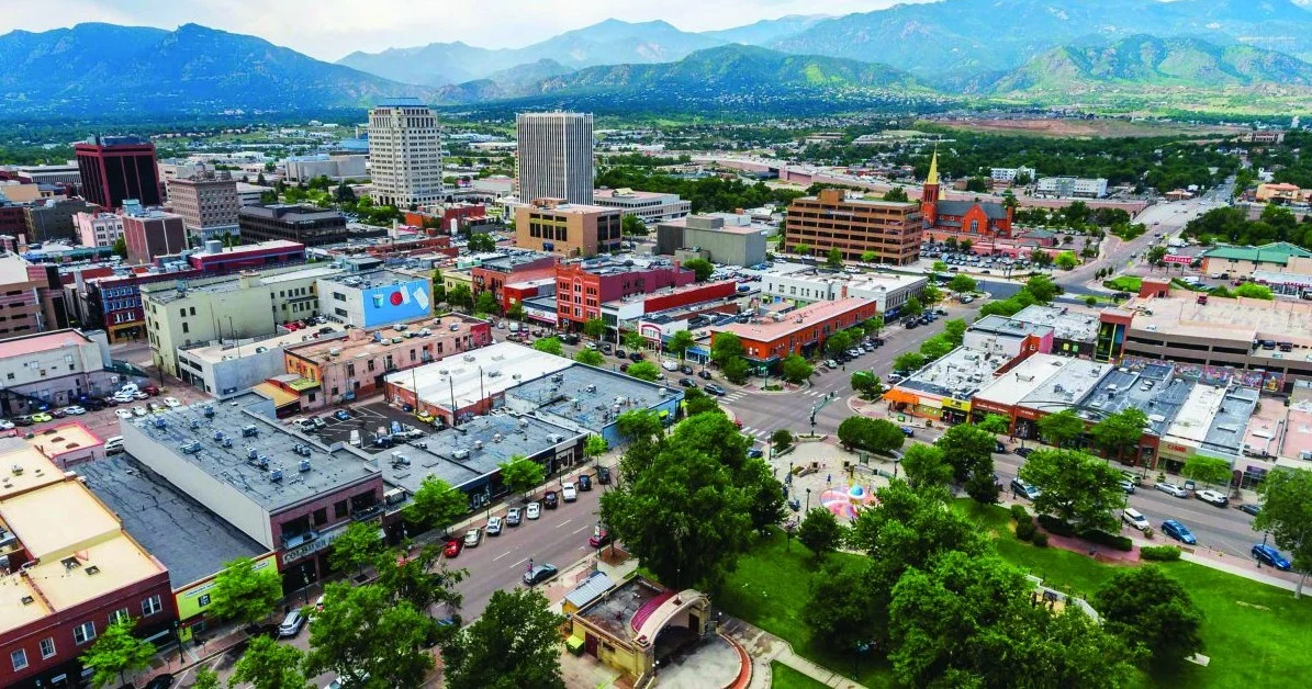 Aerial view of Colorado Springs with a mix of low-rise and high-rise buildings, tree-lined streets, mountains in the background, and bustling roads where drivers might be headed for an oil change or engine service.