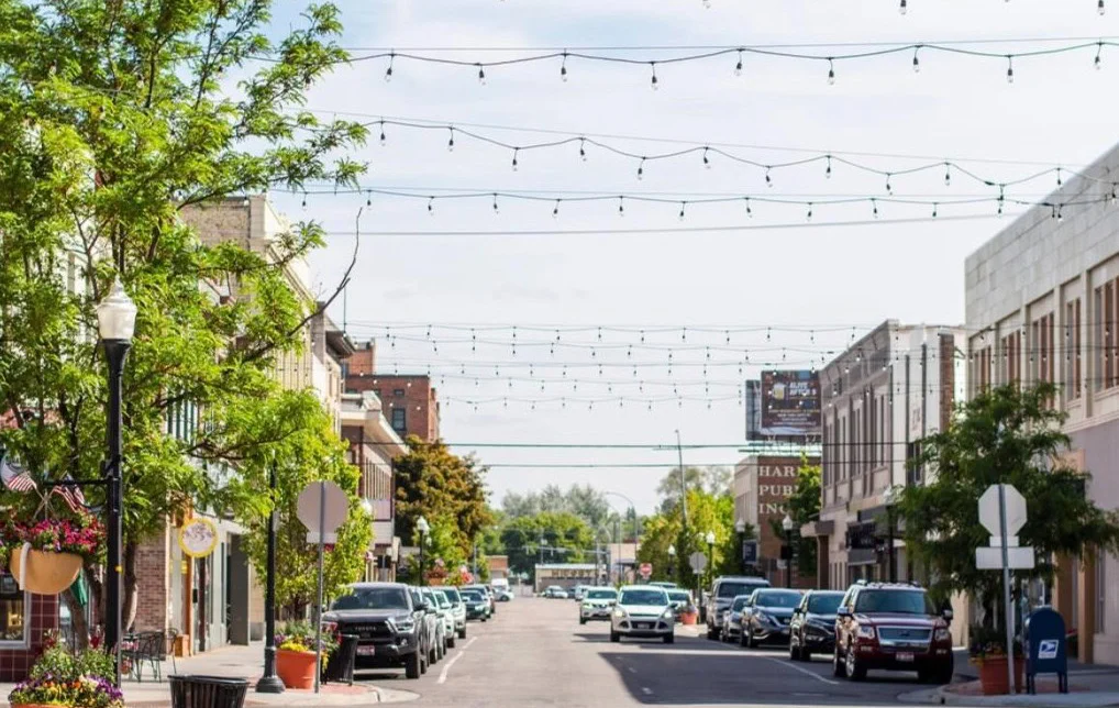 A downtown street with cars parked on both sides, string lights overhead, trees, and businesses lining the sidewalks on a sunny day.