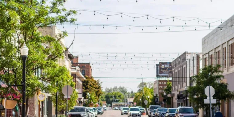 A downtown street with cars parked on both sides, string lights overhead, trees, and businesses lining the sidewalks on a sunny day.