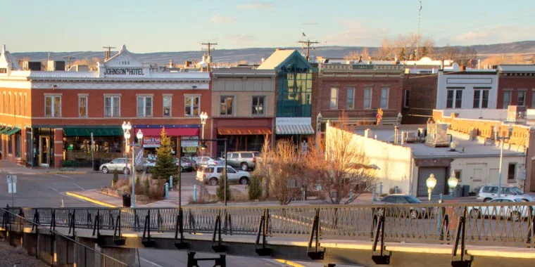 View of a small town main street with historic brick buildings, parked cars—some awaiting engine maintenance or a regular oil change—and a footbridge in the foreground under a clear sky.