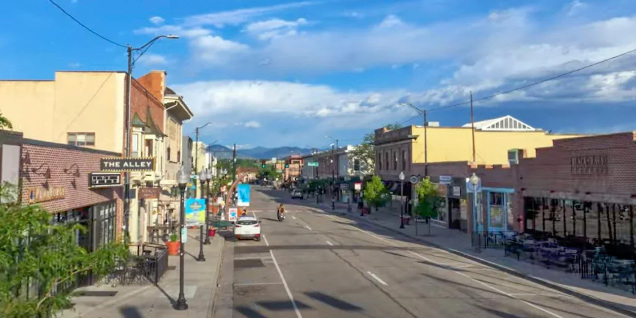 A quiet, sunlit small town street in Littleton with shops, restaurants, and few people; mountains and blue sky with clouds in the background—ideal for a peaceful walk or checking your engine's regular oil change schedule.