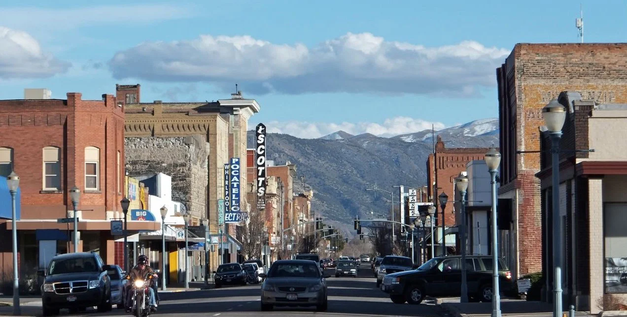 A street view of a town with brick buildings, mountains in the background, and cars driving on the road.