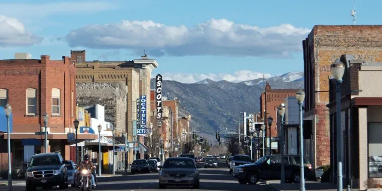 A street view of a town with brick buildings, mountains in the background, and cars driving on the road.