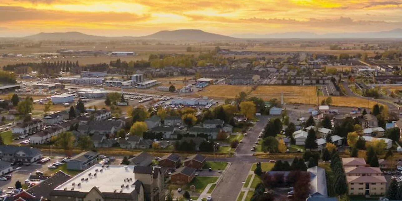A small town with residential houses and buildings is shown at sunset, with mountains and open fields visible in the background under a partly cloudy sky.