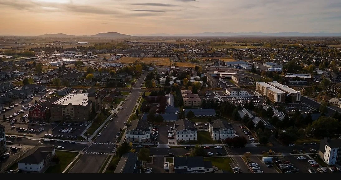 Aerial view of a suburban neighborhood with apartment buildings, houses, and parking lots. Mountains are visible in the distance under a cloudy sky.