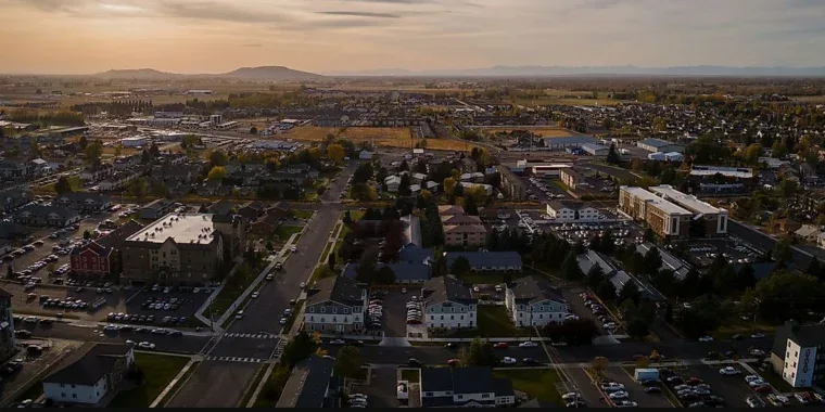 Aerial view of a suburban neighborhood with apartment buildings, houses, and parking lots. Mountains are visible in the distance under a cloudy sky.
