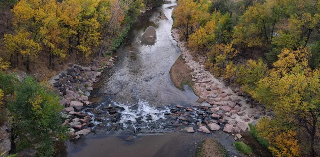A narrow river flows through a rocky, tree-lined area near Colorado Springs, with autumn foliage in shades of green, yellow, and orange.