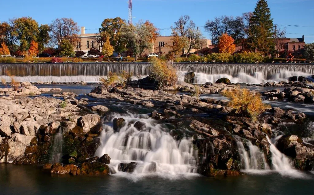 A river flows over rocky ledges and small waterfalls, with autumn foliage and buildings—home to Idaho Falls engines and oil change shops—visible in the background.