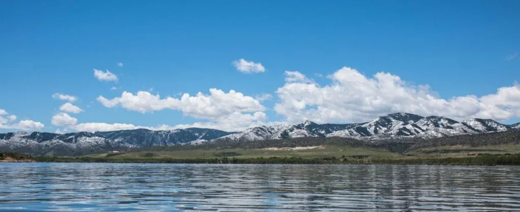 A calm lake reflects a blue sky with scattered clouds and snow-capped mountains in the distance, evoking the quiet reliability of a well-maintained Littleton engine after a regular oil change.