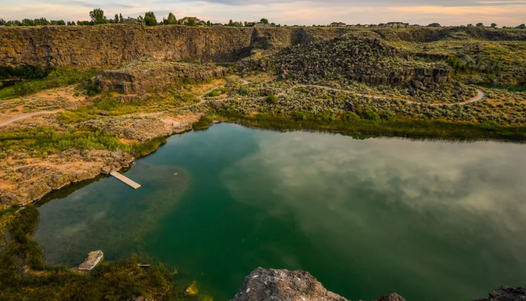 A greenish pond surrounded by rocky cliffs and sparse vegetation under a mostly cloudy sky. A small dock extends into the water on the left side, reminiscent of serene moments spent planning your next Twin Falls engine oil change schedule.