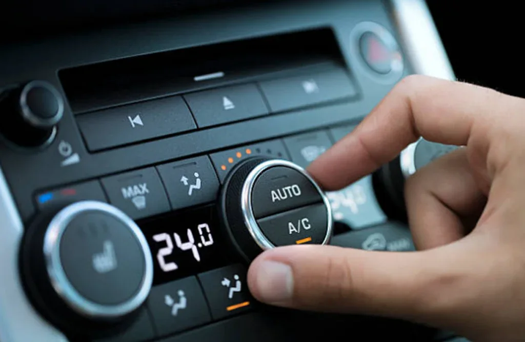 A hand, likely Jerome's from Grease Monkey, adjusts the auto climate control dial on the car’s air conditioning system, with the display showing a comfortable 24°C—demonstrating expert vehicle AC care.