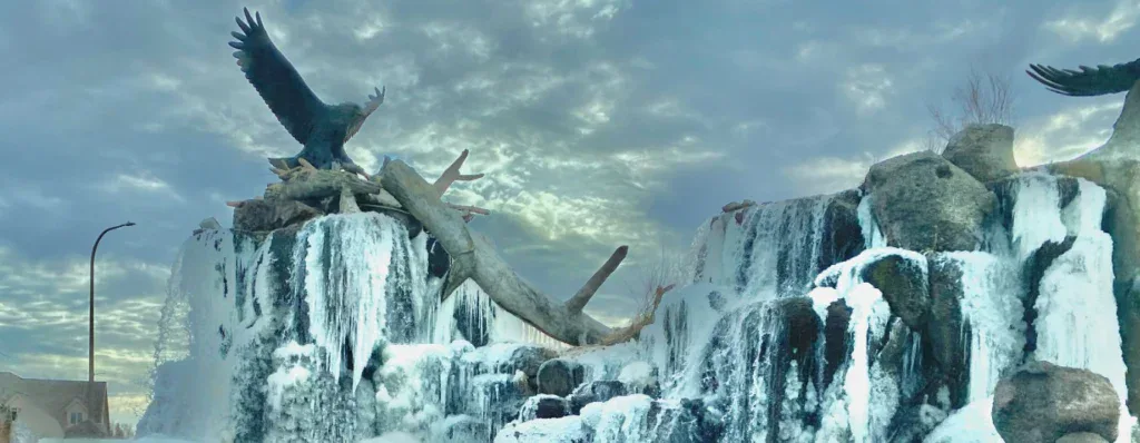 Large eagle sculptures perch on and above an icy, frozen waterfall near Idaho Falls, with rocks and a cloudy sky in the background.