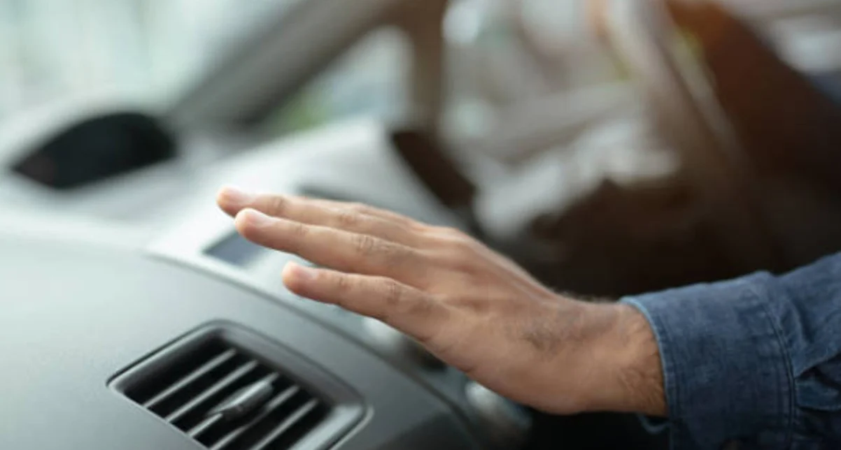 A person’s hand is held over a car air vent on the dashboard, with the driver’s hand on the steering wheel in the background—demonstrating Chevy AC Care for a perfectly cool ride.