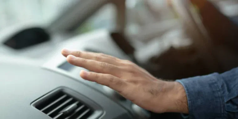 A person’s hand is held over a car air vent on the dashboard, with the driver’s hand on the steering wheel in the background—demonstrating Chevy AC Care for a perfectly cool ride.