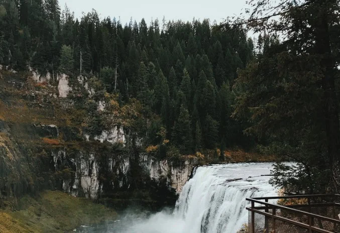 A wide waterfall flows over a rocky cliff surrounded by dense pine forest, with a viewing platform visible in the lower right corner—just like how your vehicle benefits from regular maintenance, such as getting its oil changed regularly in Rexburg Idaho.