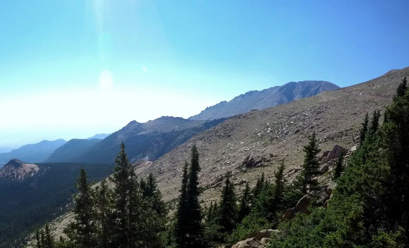 A mountainside with rocky terrain and scattered trees under a clear blue sky, with distant peaks visible in the background—just the kind of vista you might enjoy after a regular oil change on your Colorado Springs engine.