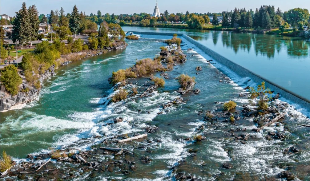 A river flows over rocks and a low dam, with trees lining the banks and a tall building visible in the background under a blue sky—Idaho Falls engines hum softly nearby.