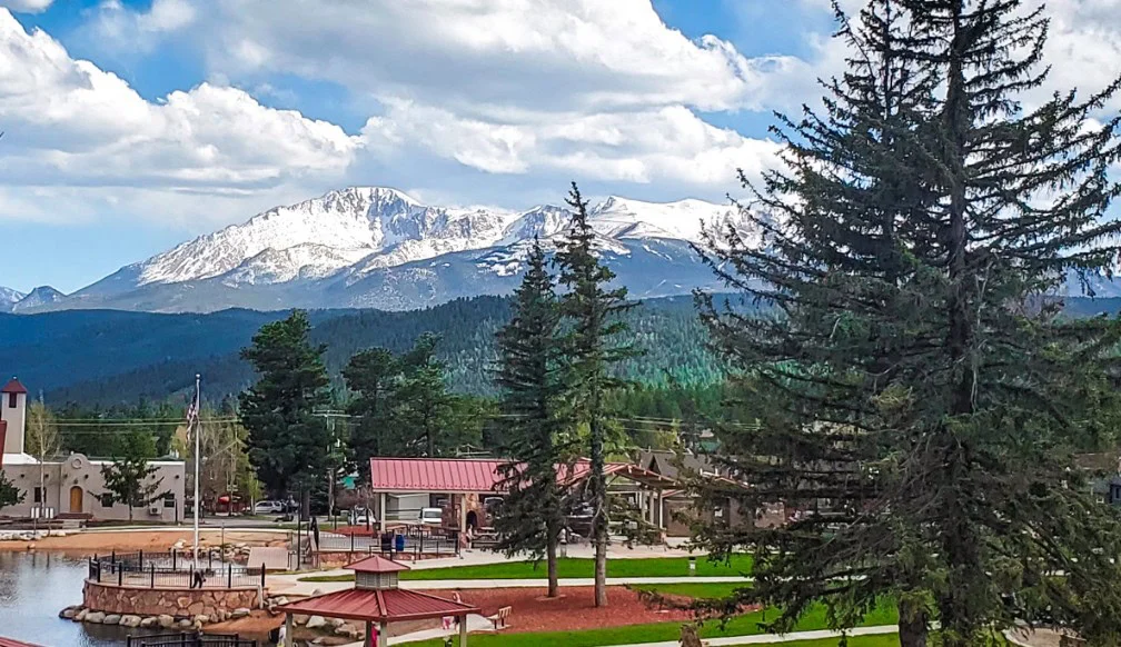 Snow-capped mountains rise behind pine trees and a grassy park, where a small building sits in the foreground—perfect for a peaceful stroll or checking your woodland park engine’s oil change schedule under the partly cloudy sky.
