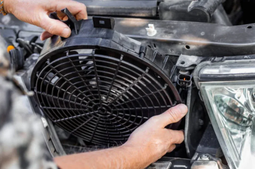 Jerome, a Grease Monkey expert, installs or removes an electric radiator fan from a car engine compartment next to a headlight as part of thorough vehicle AC care.