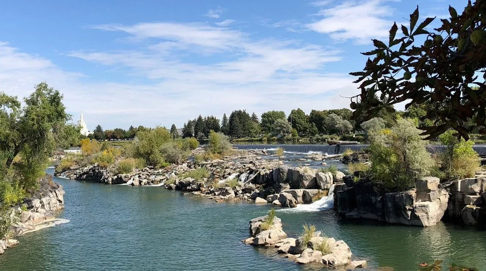 A river flows over rocks and small waterfalls near Idaho Falls, surrounded by trees and shrubs under a partly cloudy sky.