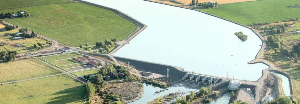 Aerial view of a dam with a reservoir near Idaho Falls, surrounded by green fields and a hydroelectric plant releasing water into a downstream river.