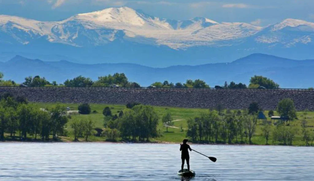 A resident paddleboards on a calm lake with trees and a dam in the background, snow-capped mountains under a cloudy sky.