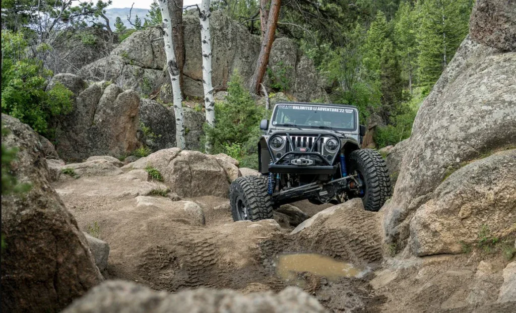 A black off-road Jeep with reliable Laramie Wyoming vehicle air conditioning navigates over large rocks and uneven terrain in a forested, mountainous area.