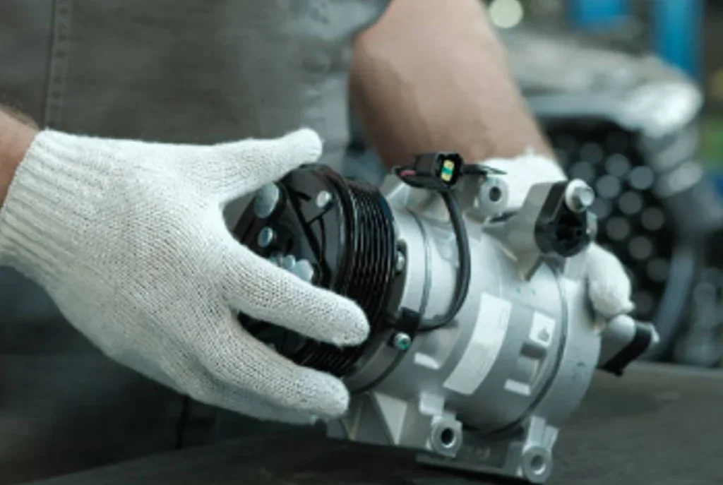 A person wearing white gloves holds a vehicle air conditioning compressor on a workbench in Idaho Falls, preparing it for installation or inspection to ensure drivers stay cool and comfortable.