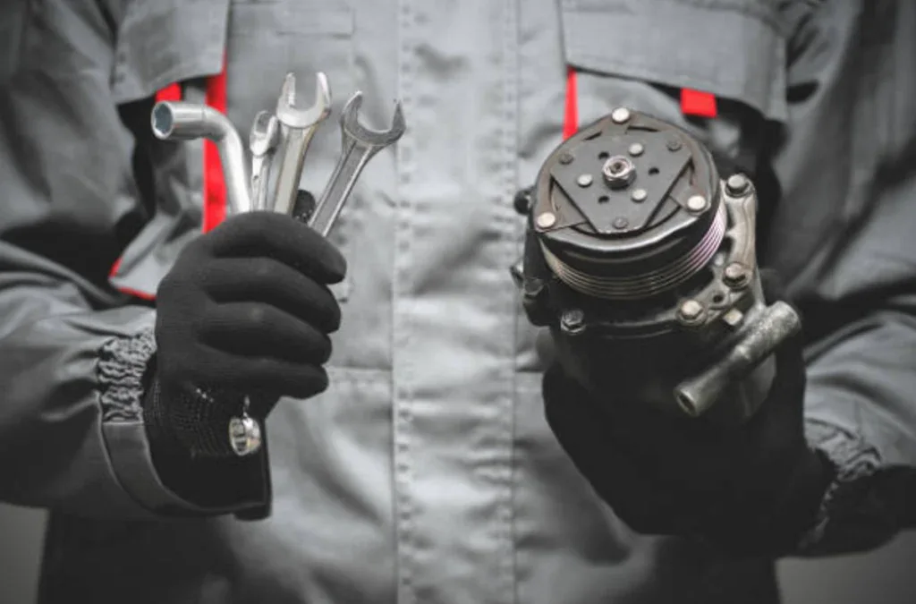 A person in gray workwear and black gloves holds wrenches in one hand and an automotive air conditioning compressor in the other, ready to provide vehicle air conditioning service for drivers in Rexburg Idaho.