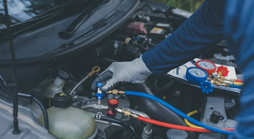 A person wearing gloves uses a gauge and hoses to check or refill a car's air conditioning system under the hood, providing essential vehicle air conditioning service for drivers in Southeast Pocatello.
