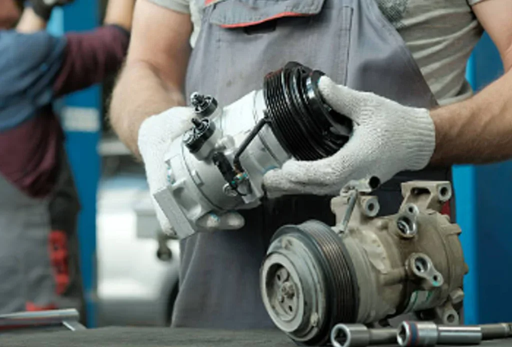 A mechanic in Idaho Falls wearing gloves holds a new automotive compressor while comparing it with an old, used compressor on a workbench during a vehicle air conditioning service.
