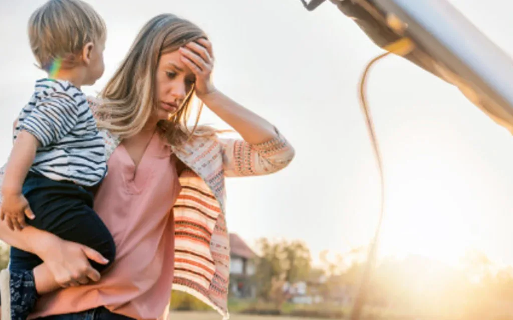 A woman holding a young child stands by a car with its hood open in Jerome, Idaho, looking concerned and touching her forehead—perhaps she’s in need of a vehicle air conditioning service or inspection.