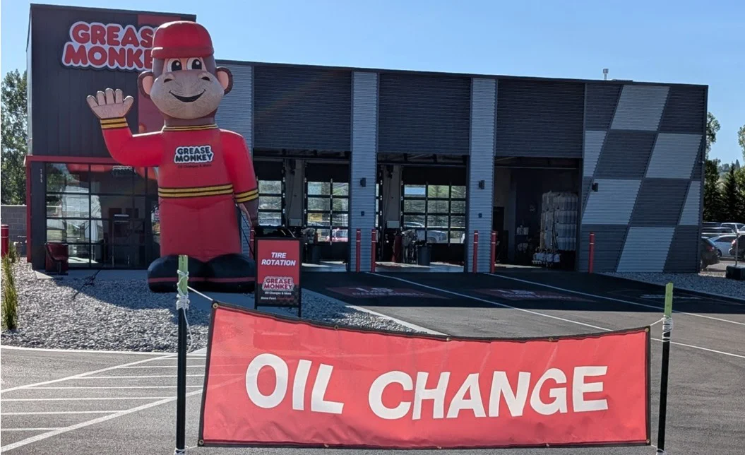 A Grease Monkey auto service center celebrates its Grand Opening, featuring a large inflatable mascot and a red banner reading "OIL CHANGE" in front of the building.